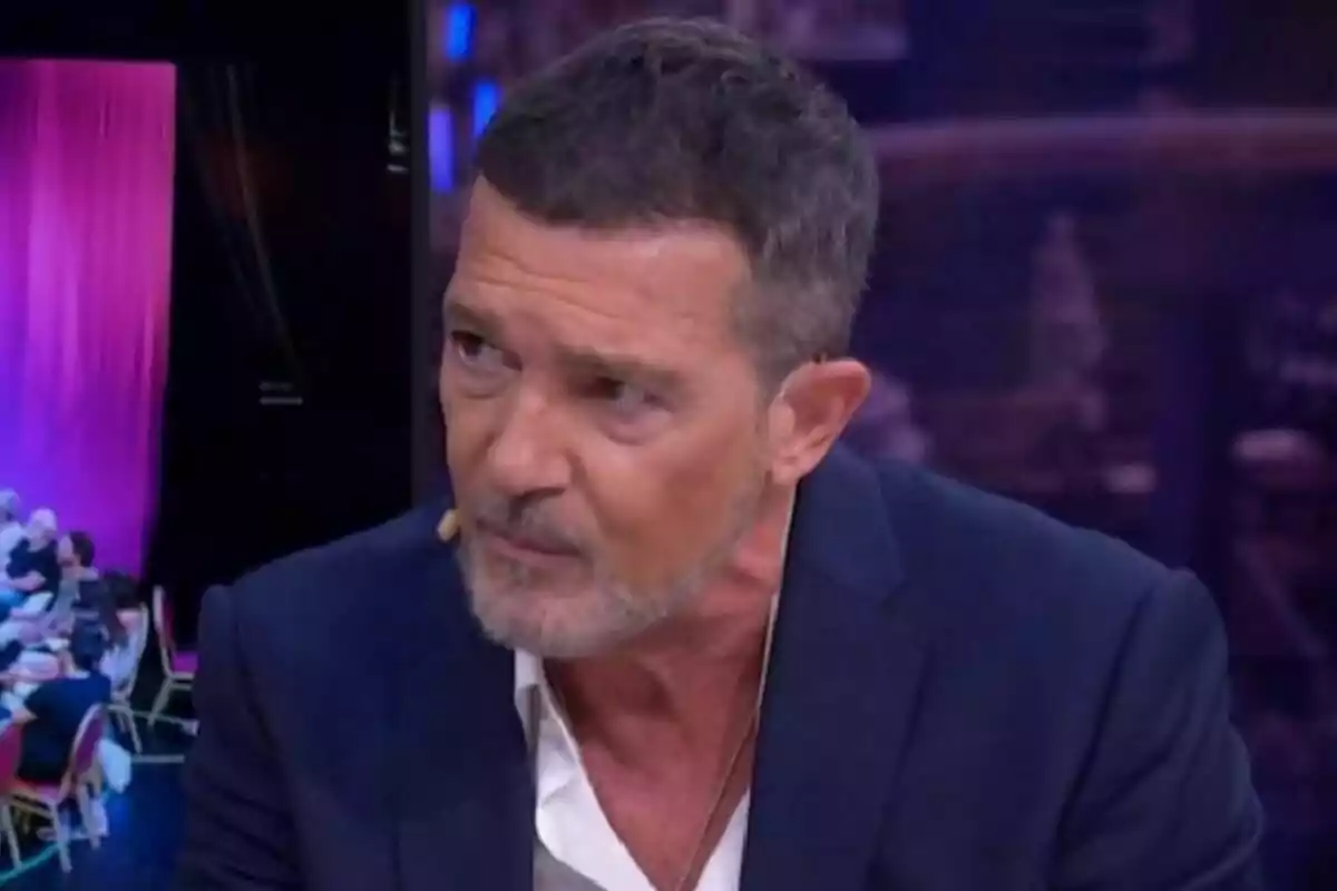 Antonio Banderas amb barba i cabell curt, vestit amb una jaqueta blava i camisa blanca, parlant a El Hormiguero amb un micròfon a l'orella.