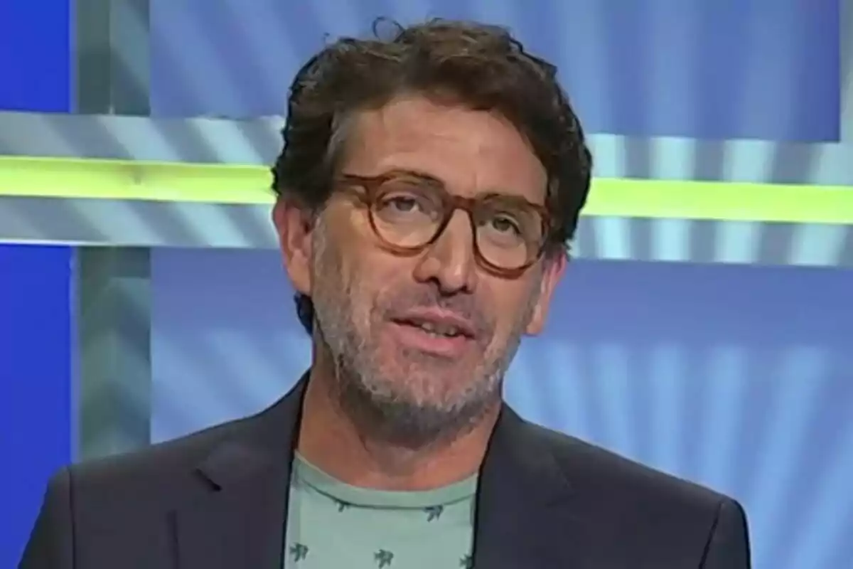 Antonio Garrido de cabell castany i ulleres marrons amb barba i samarreta verd clar, dret davant d’un fons blau amb línies grogues i grises a Atrapa’m si pots.