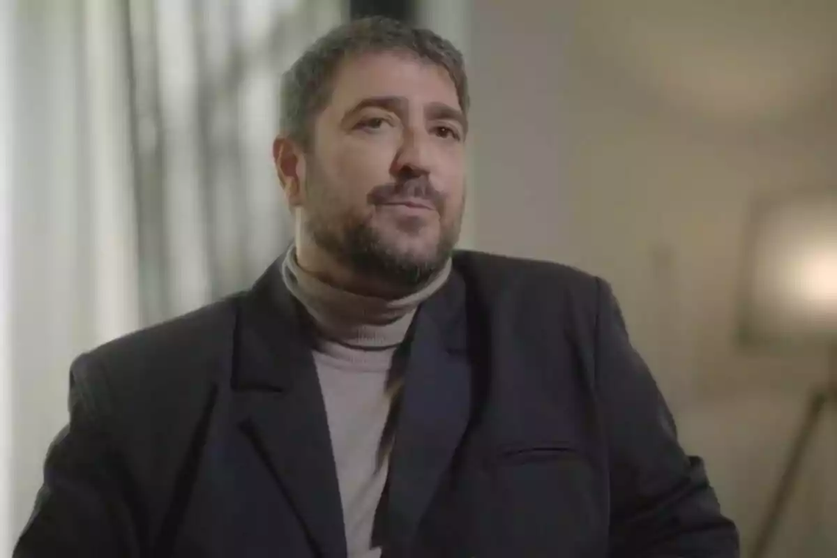 Antonio Orozco amb cabell curt i barba, vestit amb una jaqueta fosca i jersei de coll alt, assegut en un ambient interior il·luminat a 'Viatjant amb Chester'.