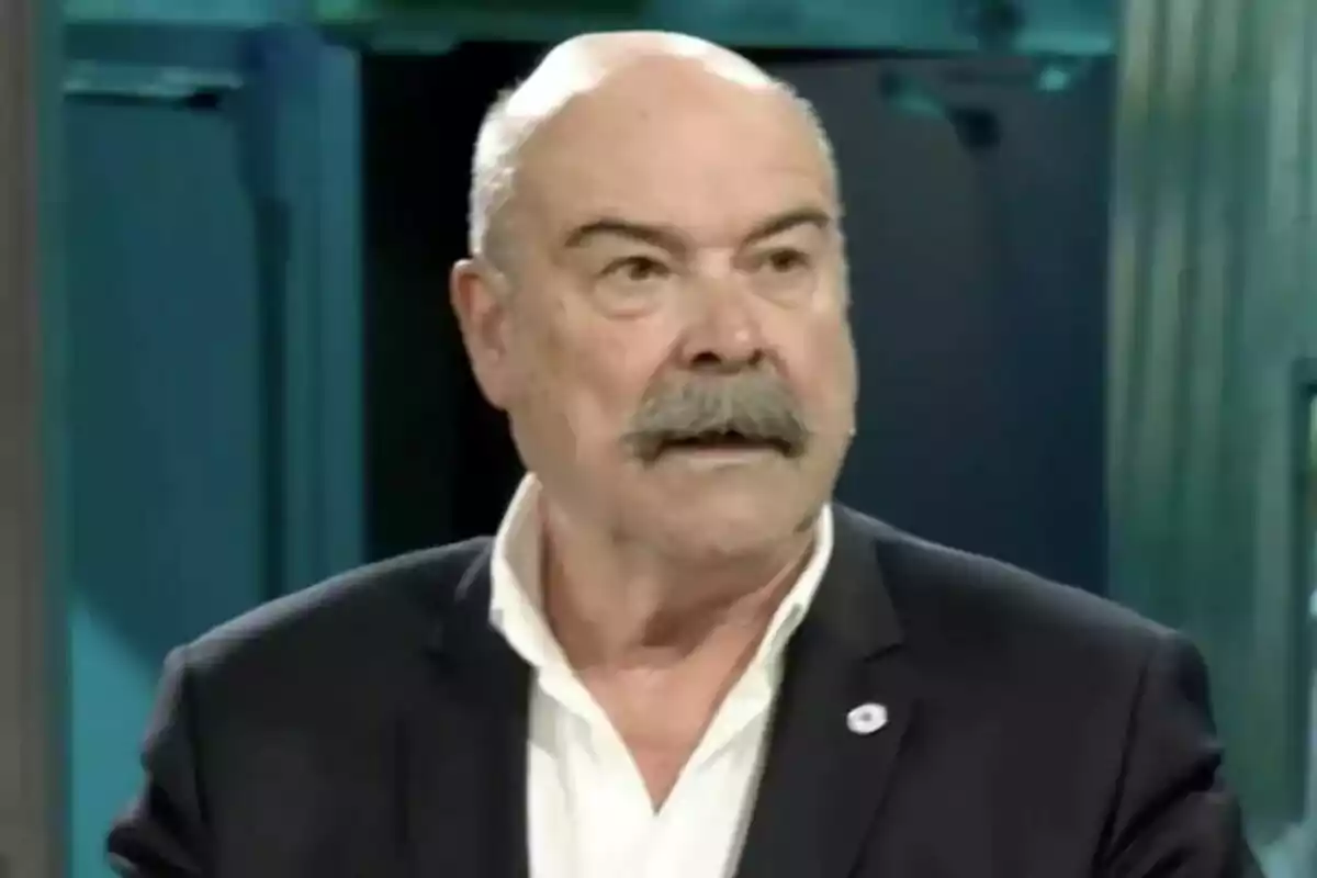 Antonio Resines amb vestit fosc i camisa blanca amb bigoti i expressió seriosa en un entorn interior a La Revuelta. Antonio Resines amb vestit fosc i camisa blanca amb bigoti i expressió seriosa en un entorn interior a La Revuelta.