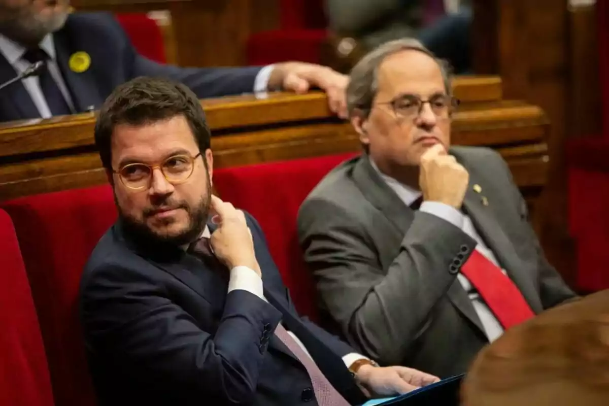 Dos homes amb vestit i corbata asseguts en un banc de fusta amb entapissat vermell en un entorn formal