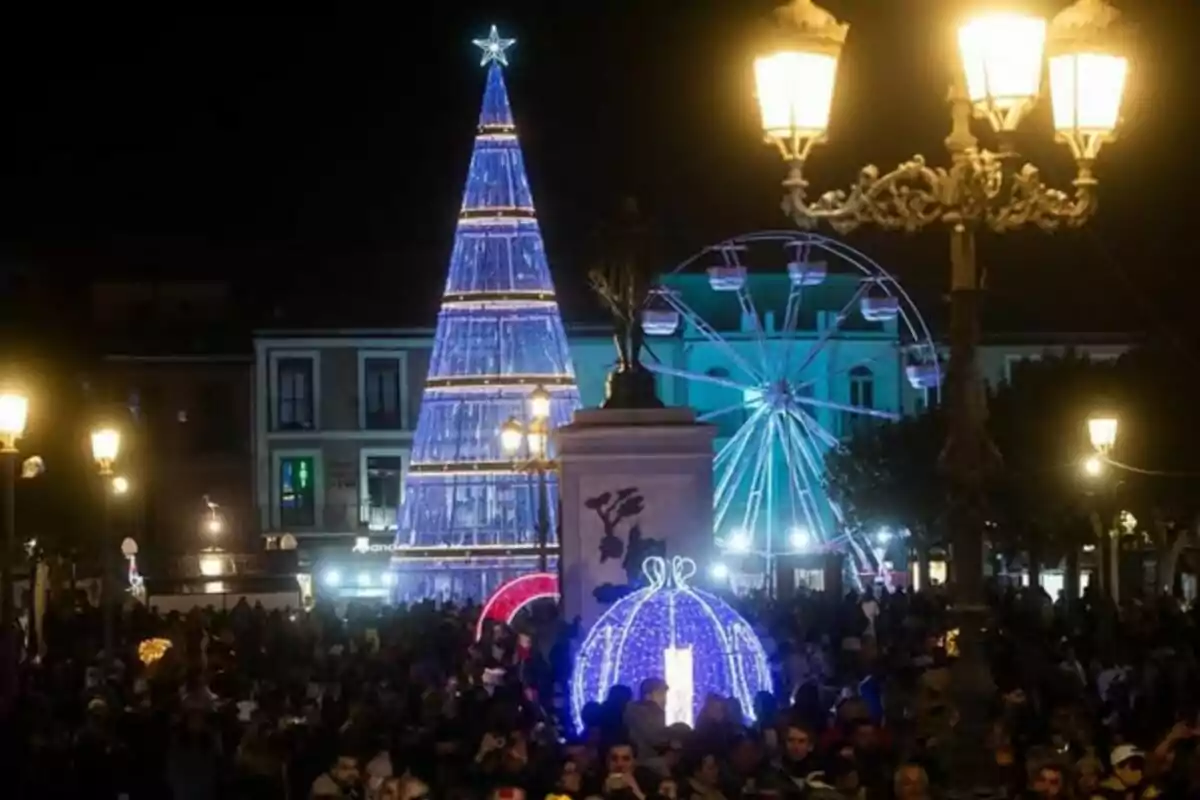 Una plaça il·luminada a la nit amb un arbre de Nadal blau, una sínia i una multitud de persones gaudint de l´ambient festiu.