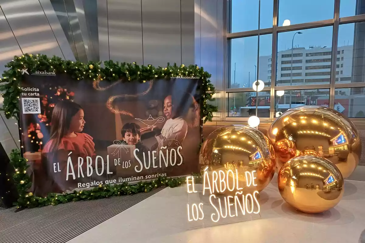 Un cartel navideño decorado con guirnaldas y luces muestra a tres niños con un regalo y el texto "El Árbol de los Sueños, Regalos que iluminan sonrisas" junto a esferas doradas brillantes y un letrero luminoso con el mismo mensaje.