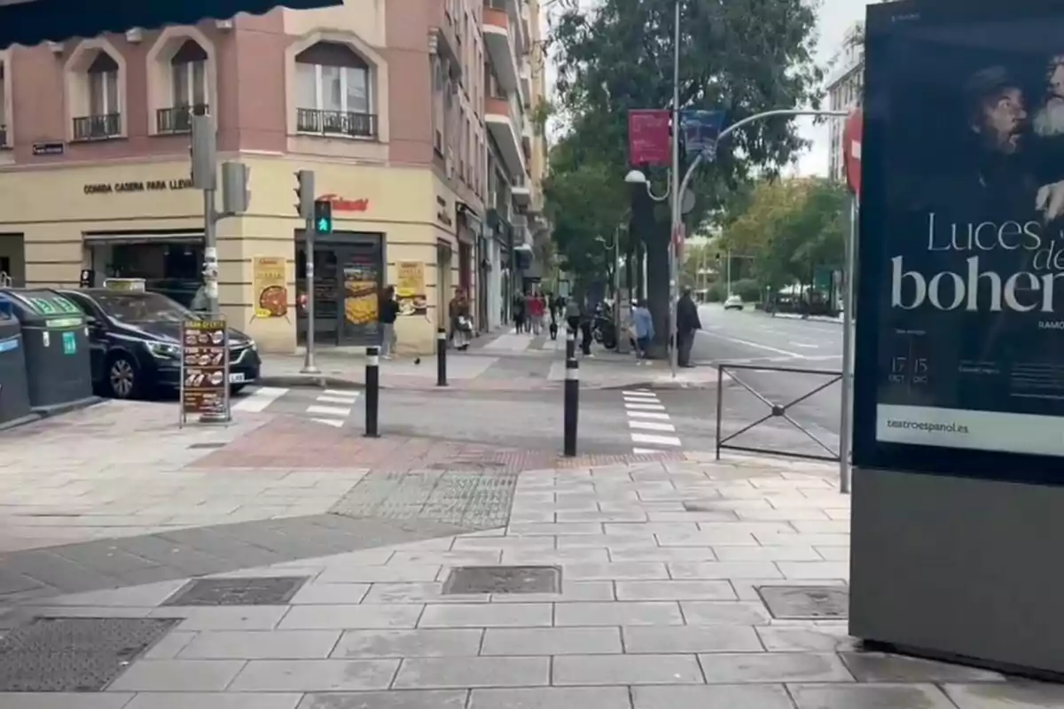 Un carrer urbà amb edificis, un semàfor en verd, un cartell publicitari i persones caminant per la vorera.