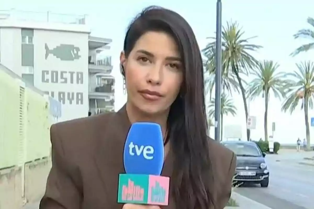 Ares Teixidó sosté un micròfon blau de La Família de la Tele amb el logotip de "tve" mentre està dempeus en un carrer amb palmeres i un edifici al fons que té un rètol que diu "Costa Platja".