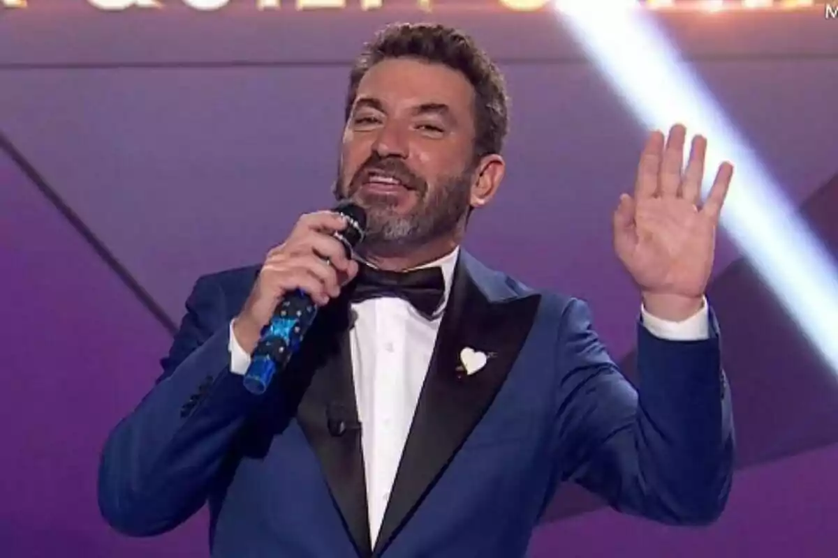 Arturo Valls amb barba i vestit blau sosté un micròfon mentre aixeca la mà al plató de Mask Singer.