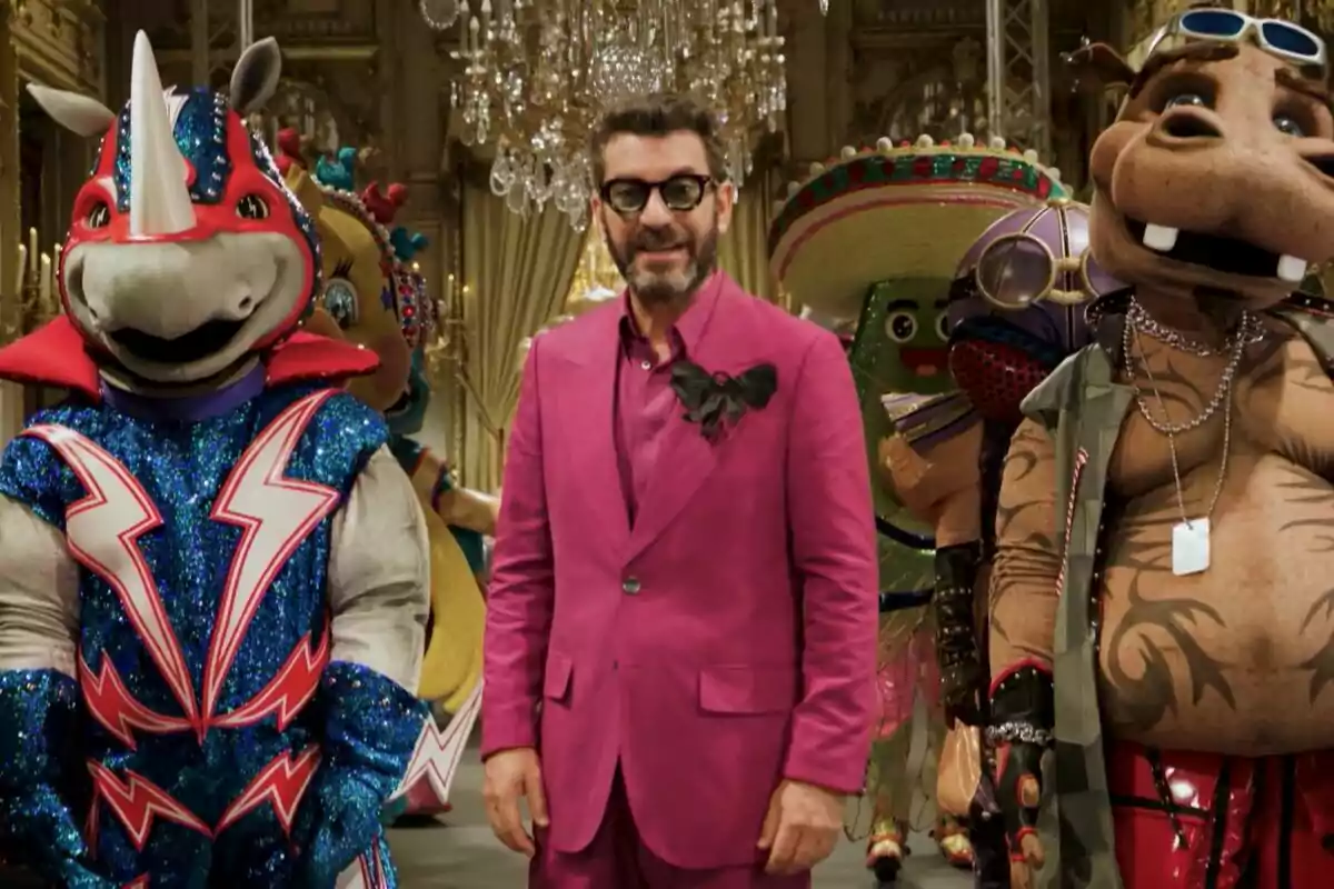 Arturo Valls amb un vestit rosa brillant està envoltat de persones disfressades d'animals i personatges colorits en un entorn elegant amb canelobres a la promo de Mask Singer.