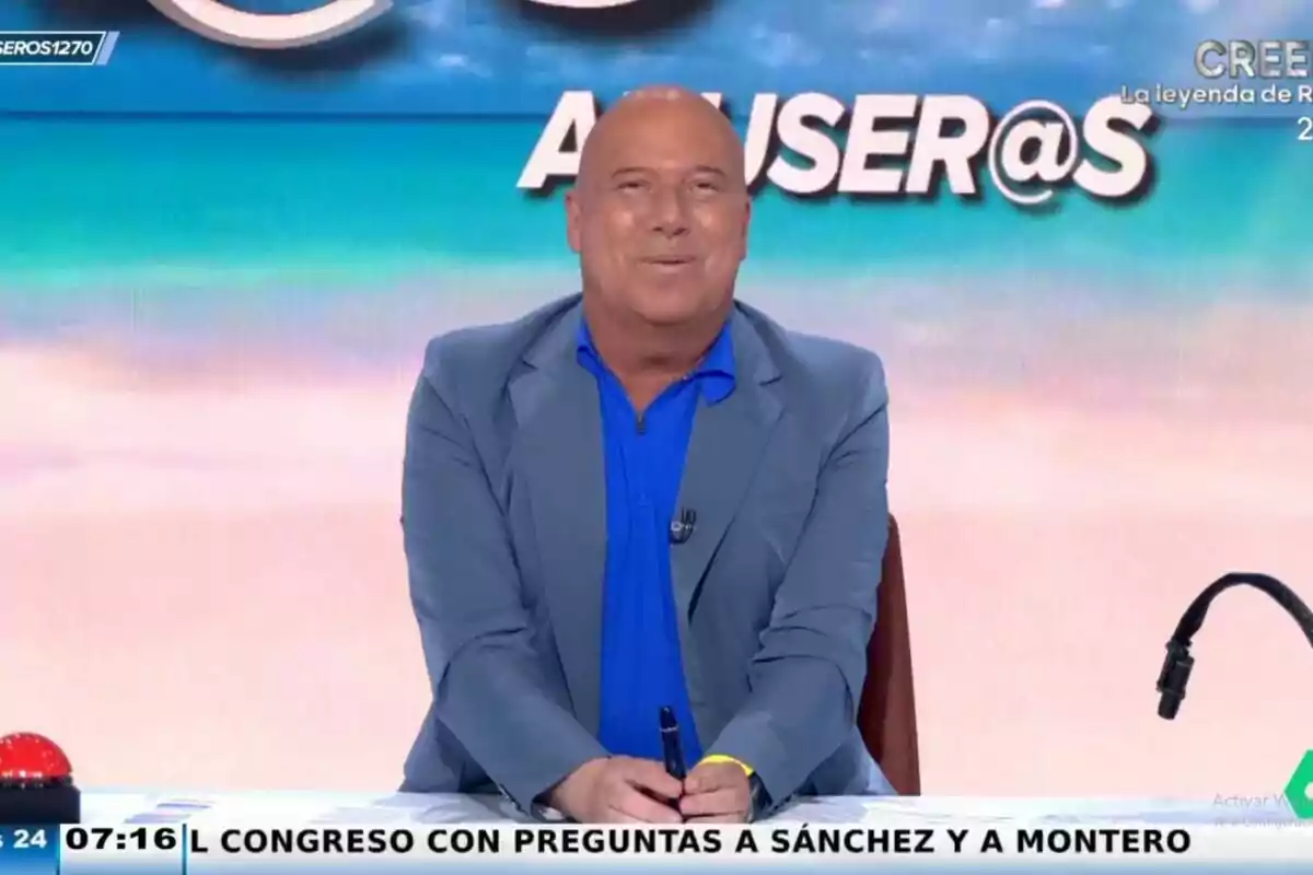 Alfonso Arús amb una jaqueta blava clar i camisa blava està assegut al set d''Aruser@s' amb un fons colorit i un rètol que diu 