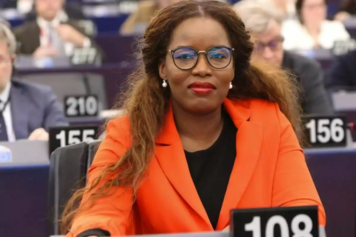 A person with glasses and an orange jacket is sitting in a conference or meeting setting, with other people and numbers visible in the background.