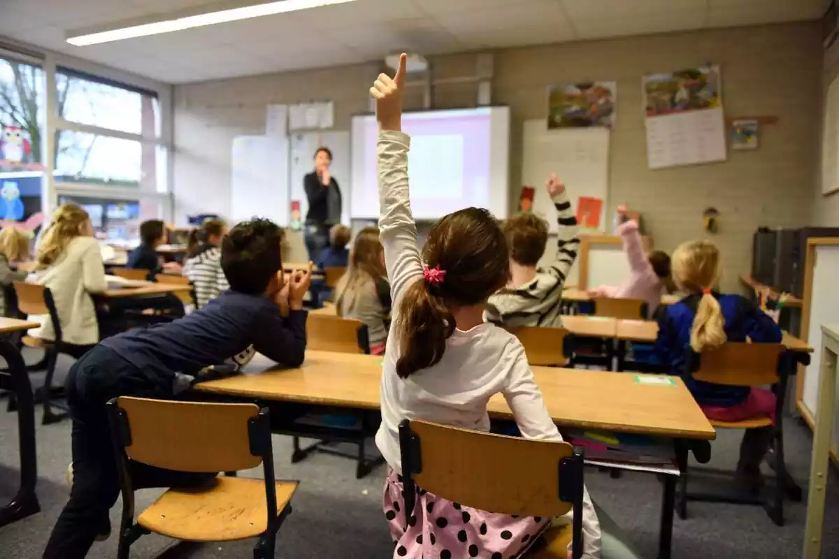 Una aula escolar vista al darrere amb diversos nens asseguts als seus pupitres i una línia aixecant el braç esquerre per preguntar a la professora que hi ha al fons