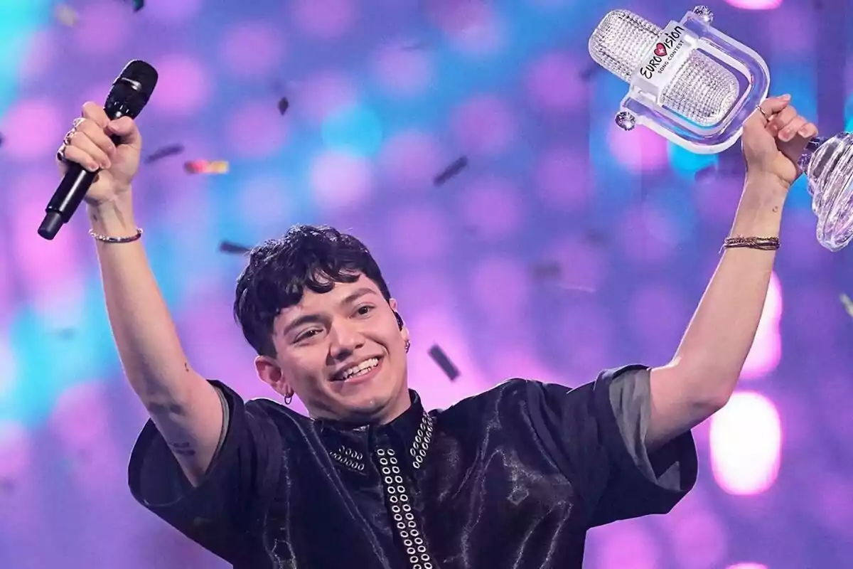 JJ somrient sosté un trofeu de micròfon de la victòria d'Eurovisió per Àustria i un micròfon en un escenari amb llums de colors i confeti.