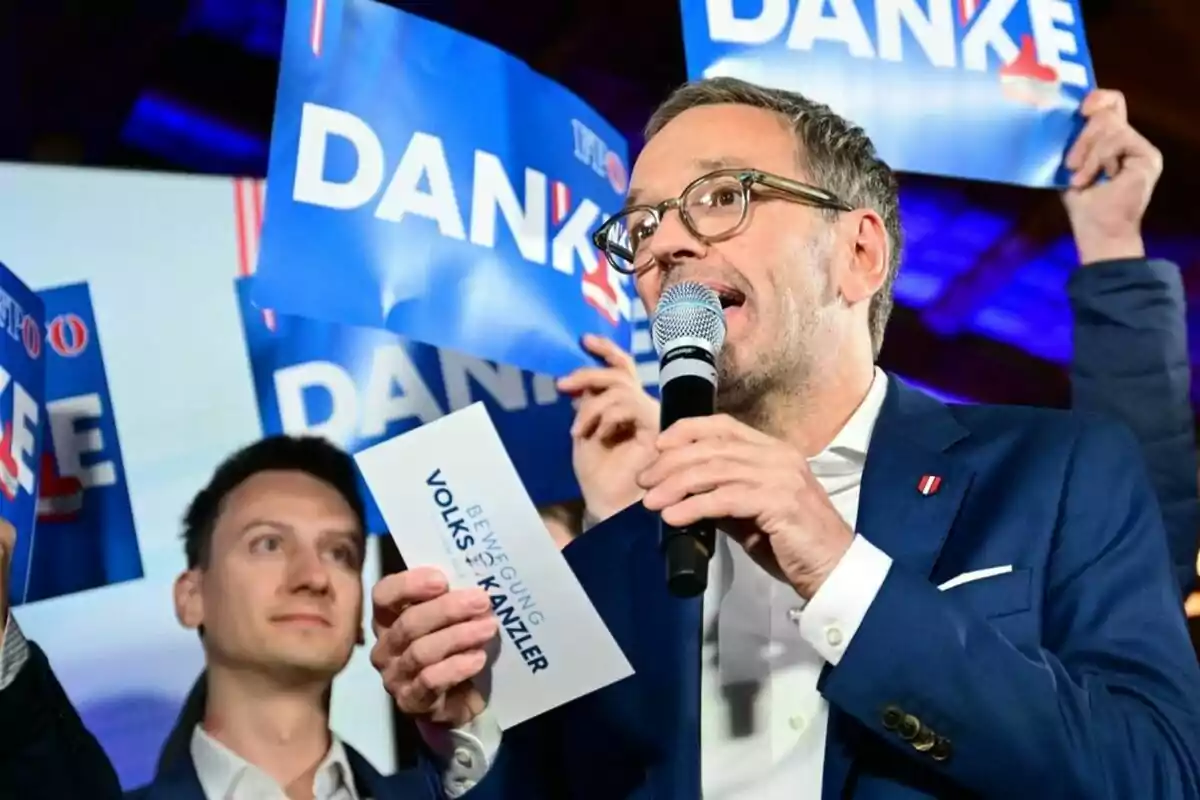 Un home amb ulleres i vestit blau sosté un micròfon mentre parla, envoltat de persones que aixequen cartells blaus amb la paraula Danke.