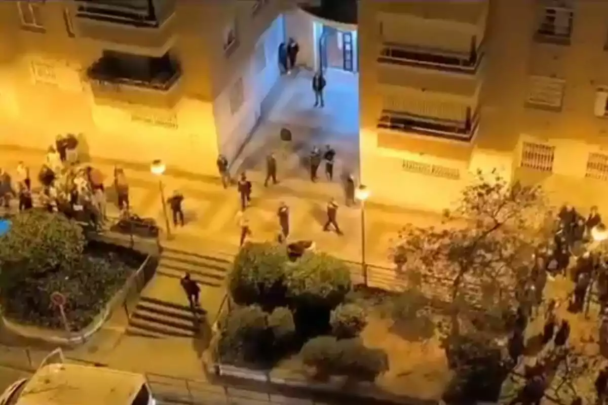 A crowd of people gathers at the entrance of a residential building at night, illuminated by streetlights.