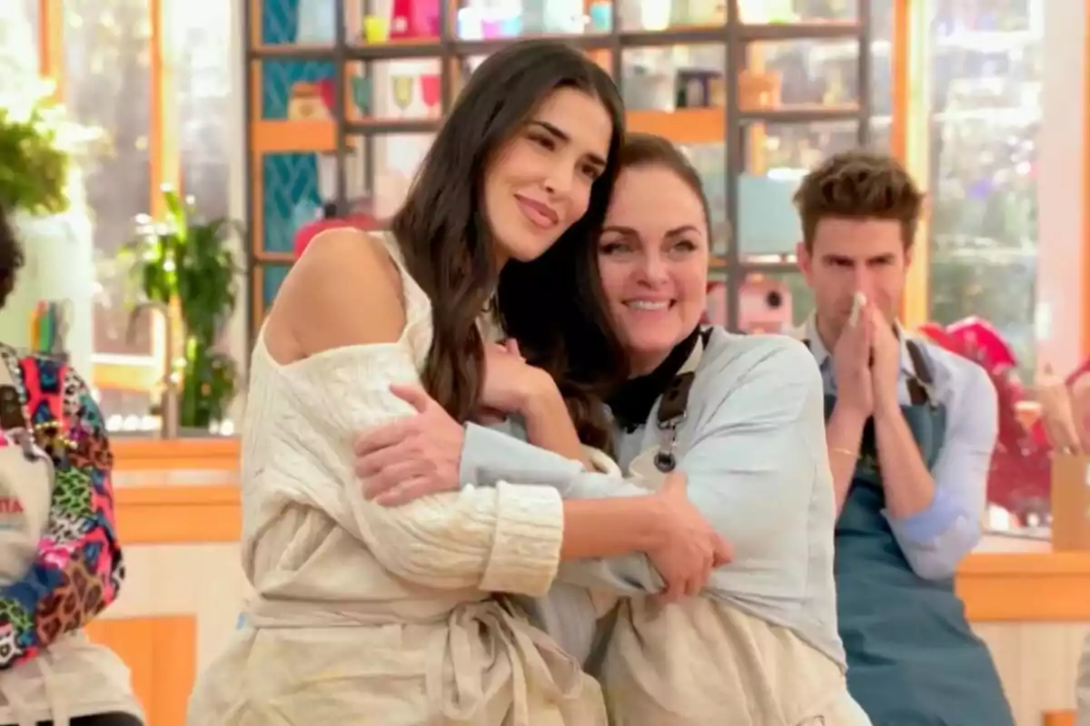 Lidia Torrent y Carmen Morales abrazándose con sonrisas en un ambiente de cocina, mientras otra persona al fondo observa con las manos juntas en 'Bake off: Famosos al horno'.