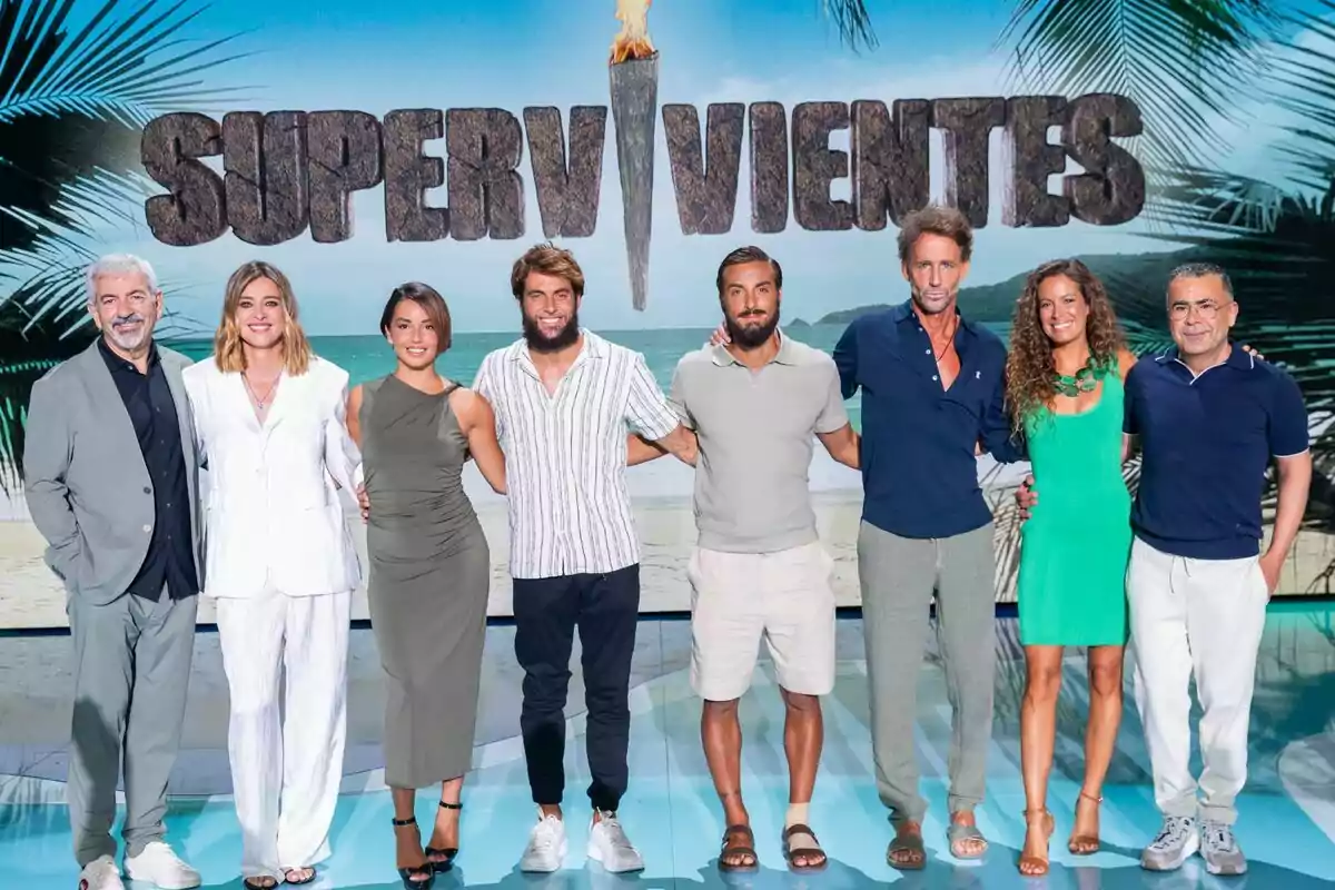 Carlos Sobera, Sandra Barneda, Anita Williams, Montoya, Borja González, Álvaro Muñoz Escassi, Laura Madrueño i Jorge Javier Vázquez posen somrients davant d’un cartell de Supervivientes amb un fons de platja i palmeres.