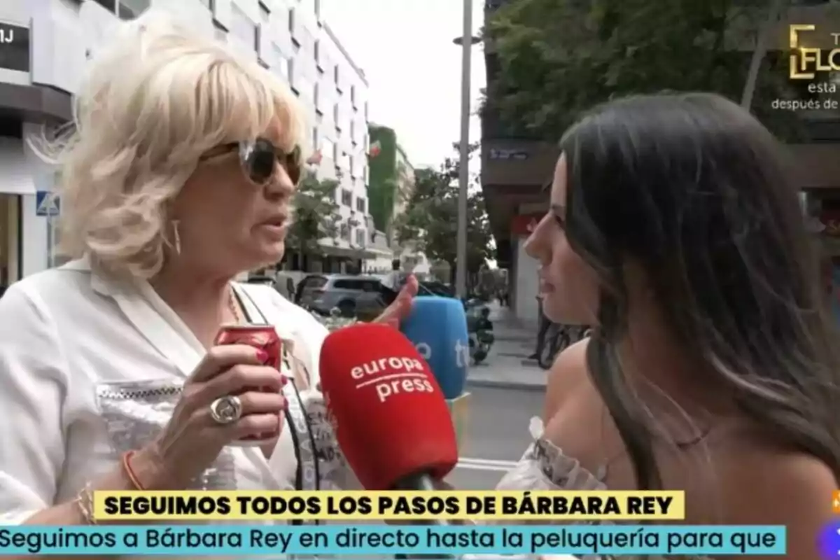 Dues dones conversen al carrer mentre Bárbara Rey sosté una llauna i diverses reporteres els acosten micròfons de diferents mitjans de comunicació com La Família de la Tele.