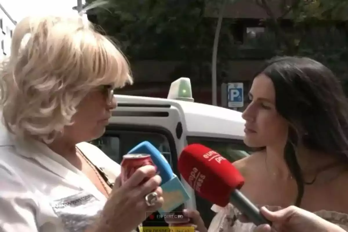Dues dones conversen al carrer mentre Bárbara Rey sosté una llauna de refresc i l’altra diversos micròfons de premsa com La Família de la Tele davant d’un taxi blanc. Dues dones conversen al carrer mentre Bárbara Rey sosté una llauna de refresc i l’altra diversos micròfons de premsa com La Família de la Tele davant d’un taxi blanc.