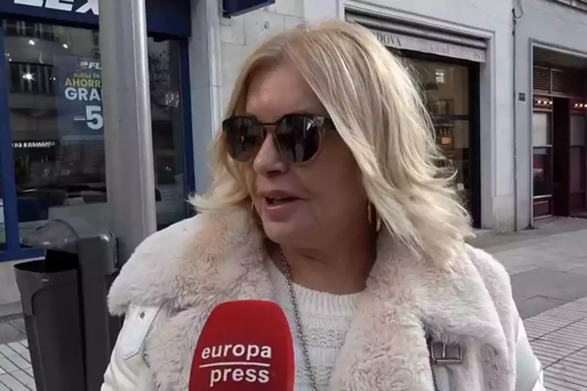 Bárbara Rey wearing sunglasses and a light coat holds a Europa Press microphone on an urban street.