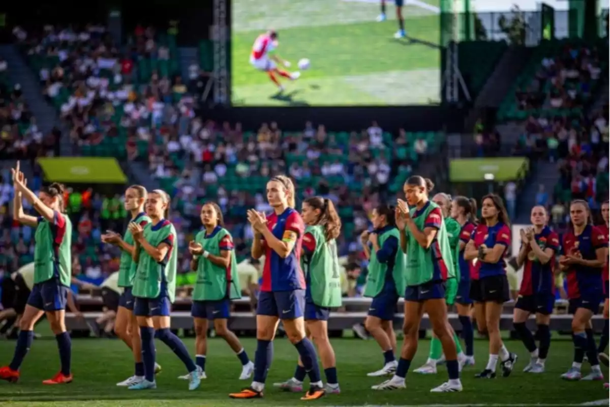 Soccer players applaud on the field while the crowd watches from the stands and a screen shows a play. Soccer players applaud on the field while the crowd watches from the stands and a screen shows a play.