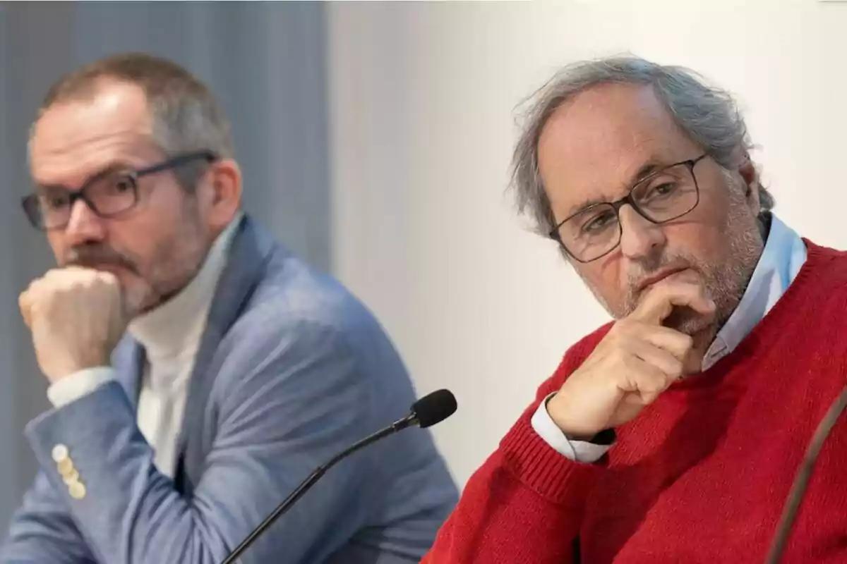 Dos homes asseguts en una taula durant una conferència, tots dos amb expressió pensativa i portant ulleres.