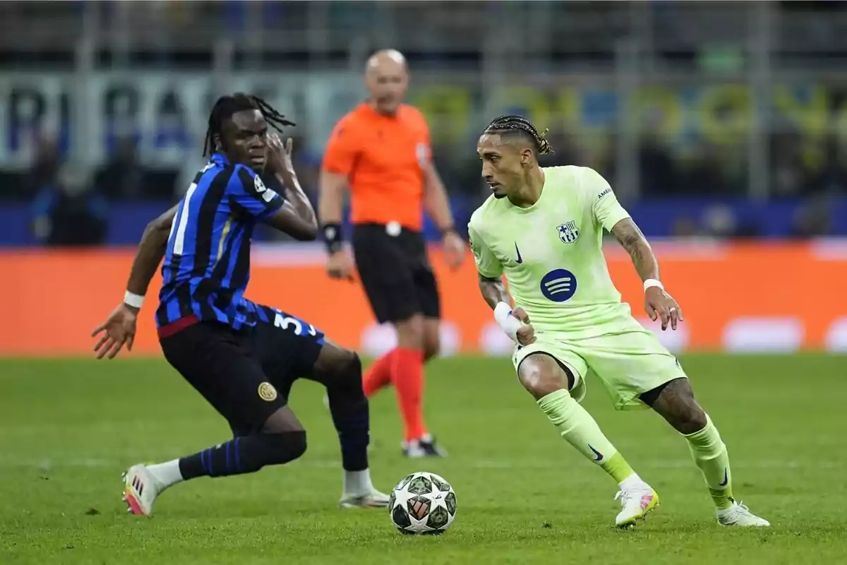 Dos jugadors de futbol competeixen per la pilota en un partit, un amb uniforme blau i negre i l'altre amb uniforme verd clar, mentre un àrbitre observa al fons. Dos jugadors de futbol competeixen per la pilota en un partit, un amb uniforme blau i negre i l'altre amb uniforme verd clar, mentre un àrbitre observa al fons.
