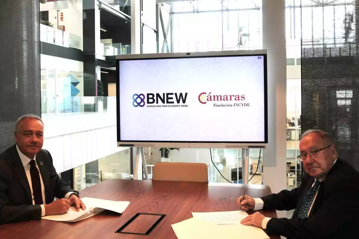 Dos càrrecs de la Fundació INCYDE i de la BNEW asseguts en una taula signant uns papers amb una pantalla de fons