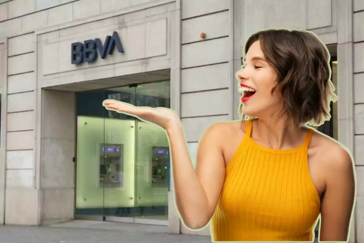 Una dona somrient amb un vestit groc sosté la mà estesa davant d'una sucursal del banc BBVA.