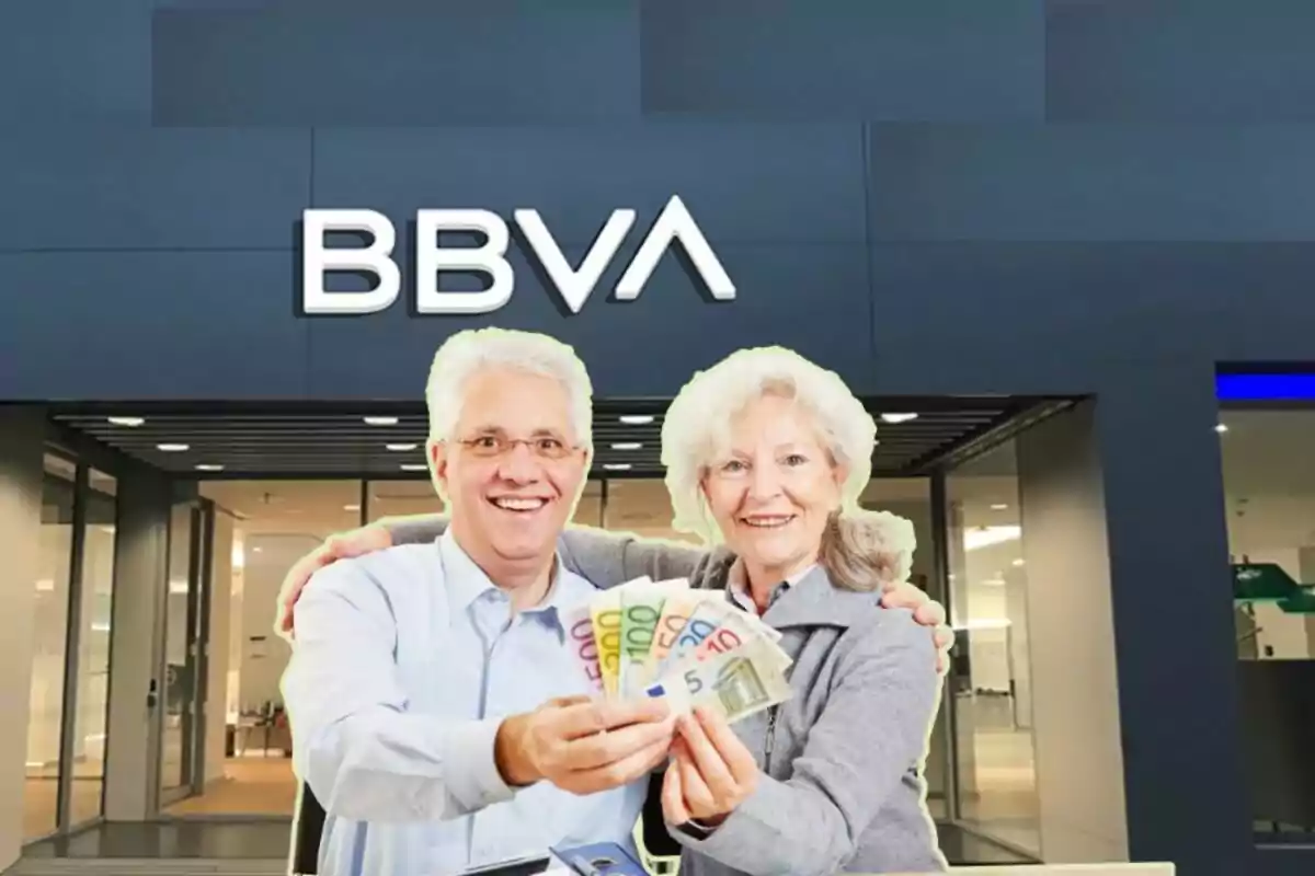 Dues persones grans somrients sostenint bitllets d'euro davant d'una sucursal del banc BBVA.