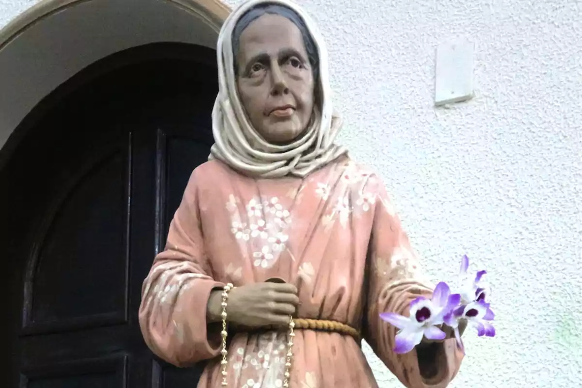 Escultura d’una dona amb vestit rosa i mocador al cap sostenint un rosari i unes flors morades davant d’una porta fosca Escultura d’una dona amb vestit rosa i mocador al cap sostenint un rosari i unes flors morades davant d’una porta fosca