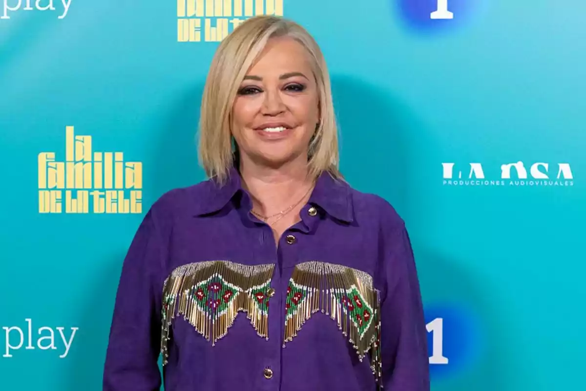 Belén Esteban amb cabell ros i camisa morada decorada, posant davant d'un fons blau amb logotips de Belén Esteban amb cabell ros i camisa morada decorada, posant davant d'un fons blau amb logotips de