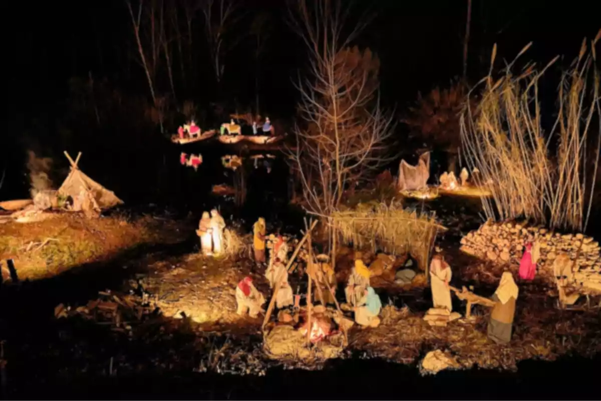 Una escena nocturna d'un pessebre vivent amb figures que representa persones al voltant d'una foguera i altres activitats en un entorn natural amb arbres i aigua.