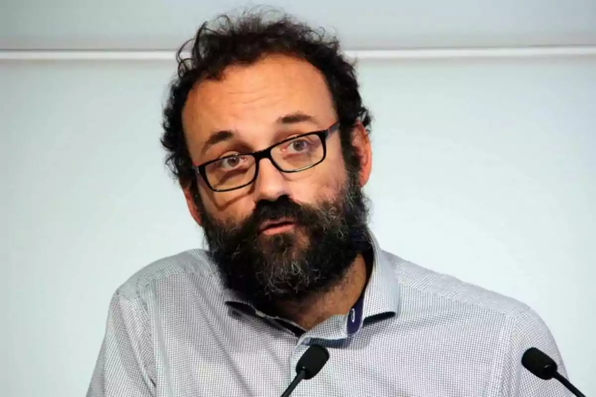 Bearded man with glasses speaking in front of two microphones, wearing a light-colored jersey and a neutral background