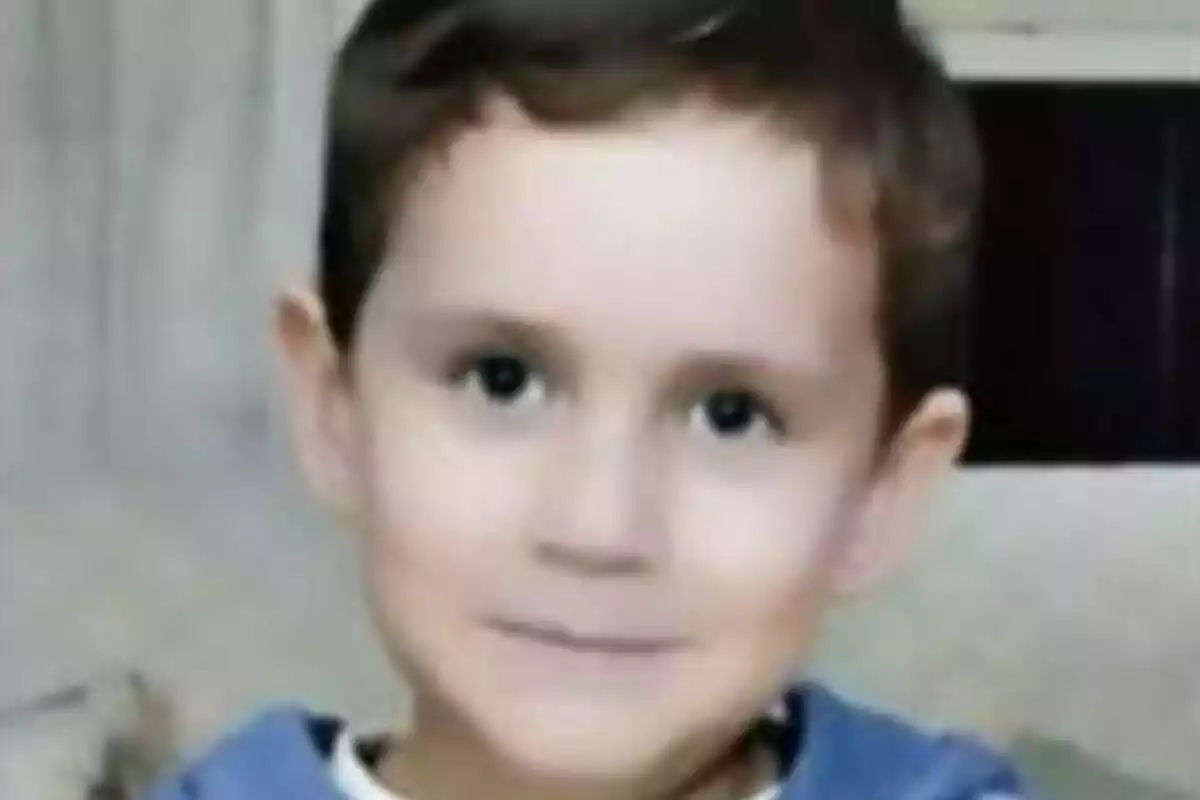 Un nen petit amb cabell castany i ulls foscos, vestint una samarreta blava, mira a la càmera amb una expressió neutral.
