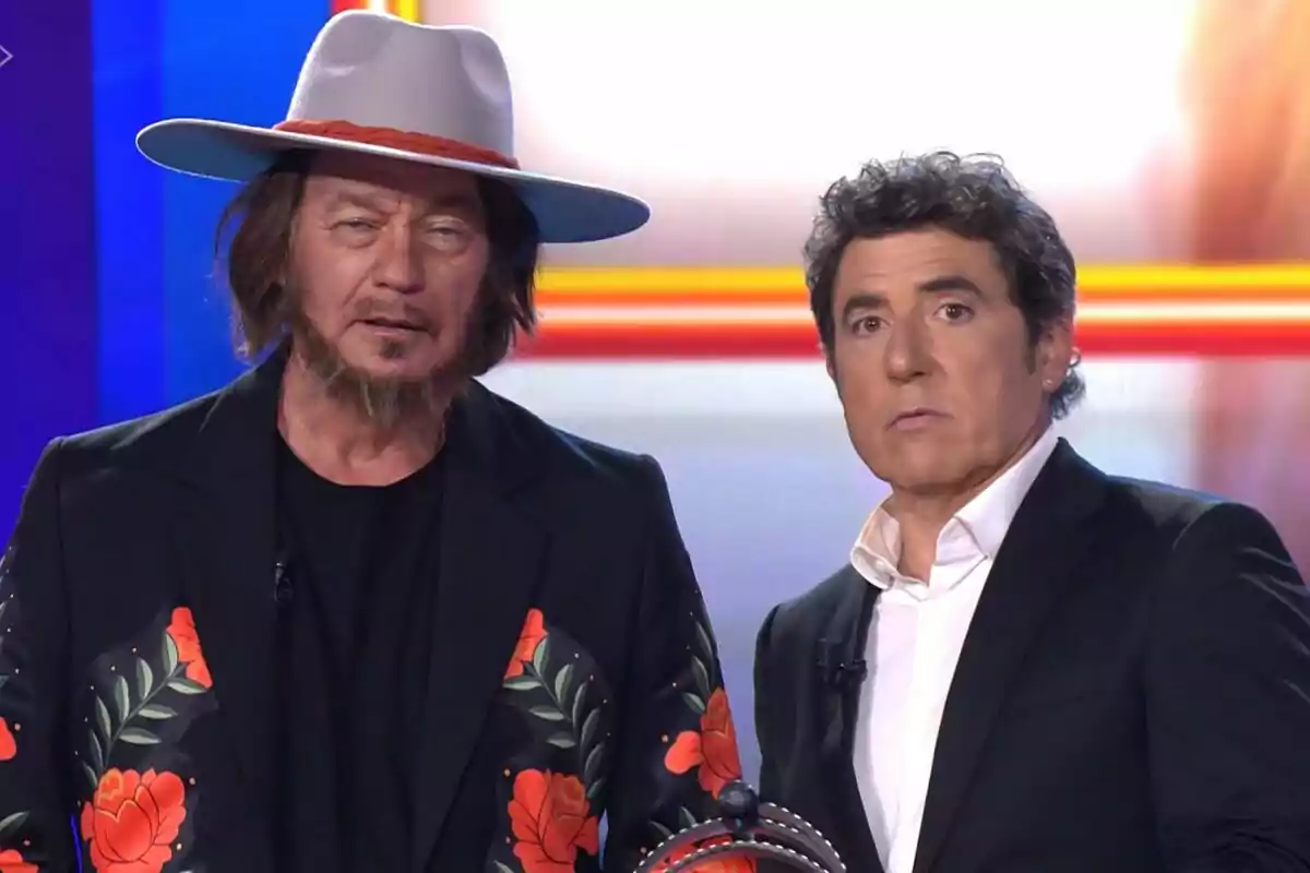 Bertín Osborne i Manel Fuentes en un plató de Tu Cara Me Suena, un porta barret gris i jaqueta amb flors vermelles i l’altre vesteix vestit negre amb camisa blanca.