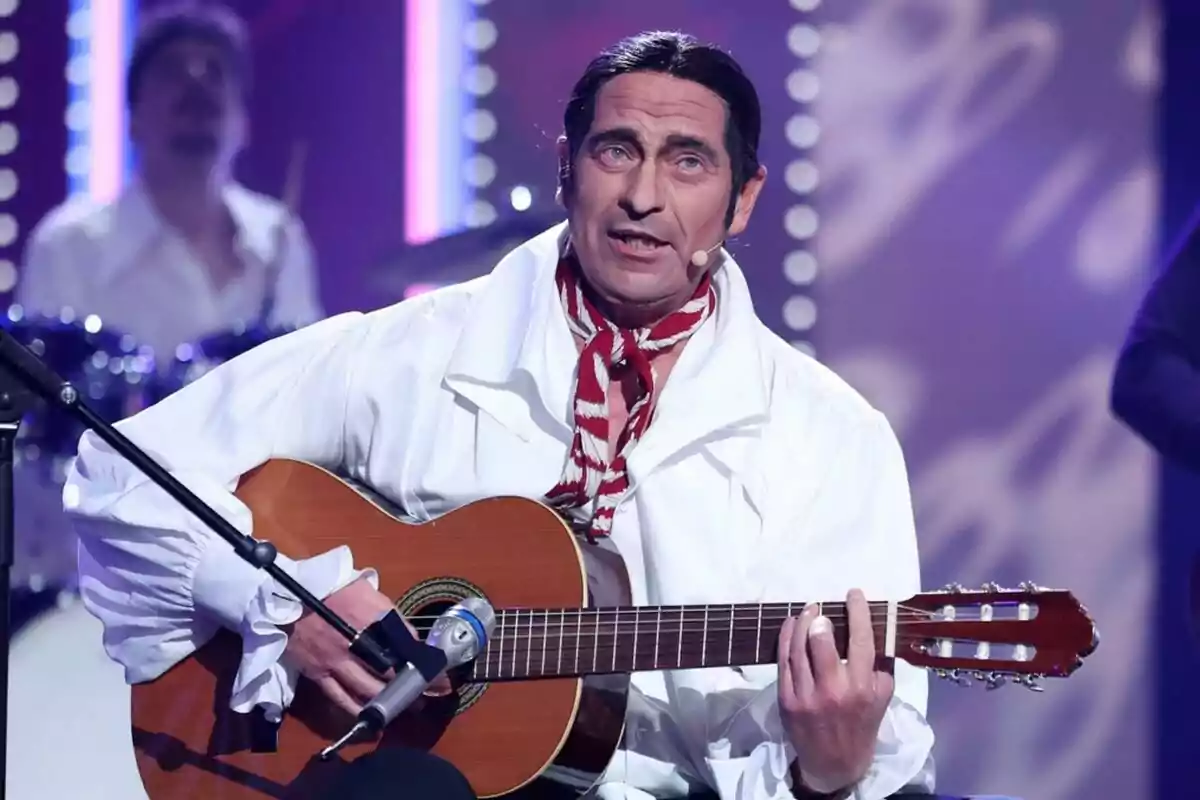 Bertín Osborne amb camisa blanca i mocador vermell toca la guitarra en un escenari il·luminat a Tu Cara Me Suena. Bertín Osborne amb camisa blanca i mocador vermell toca la guitarra en un escenari il·luminat a Tu Cara Me Suena.