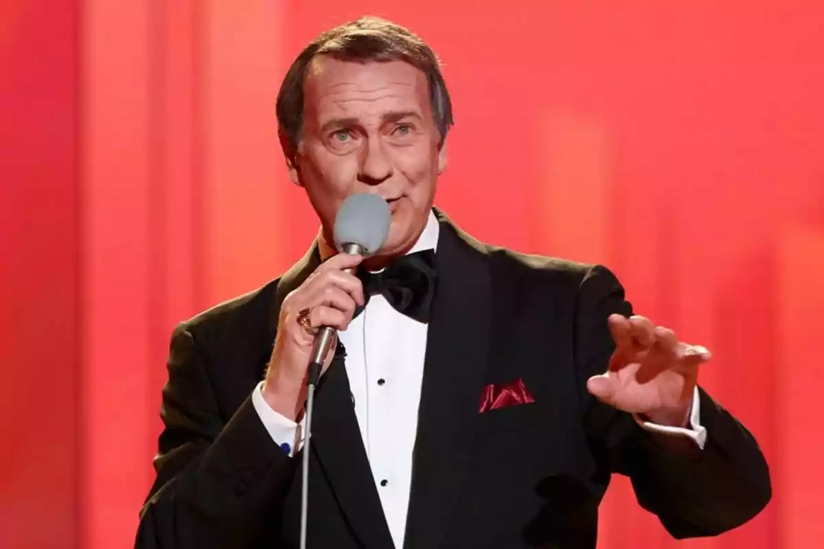 Bertín Osborne en un escenari amb un micròfon a la mà vestit d'esmòquing negre amb llacet i mocador vermell a la butxaca de l'americana fons vermell a Tu Cara Me Suena. Bertín Osborne en un escenari amb un micròfon a la mà vestit d'esmòquing negre amb llacet i mocador vermell a la butxaca de l'americana fons vermell a Tu Cara Me Suena.
