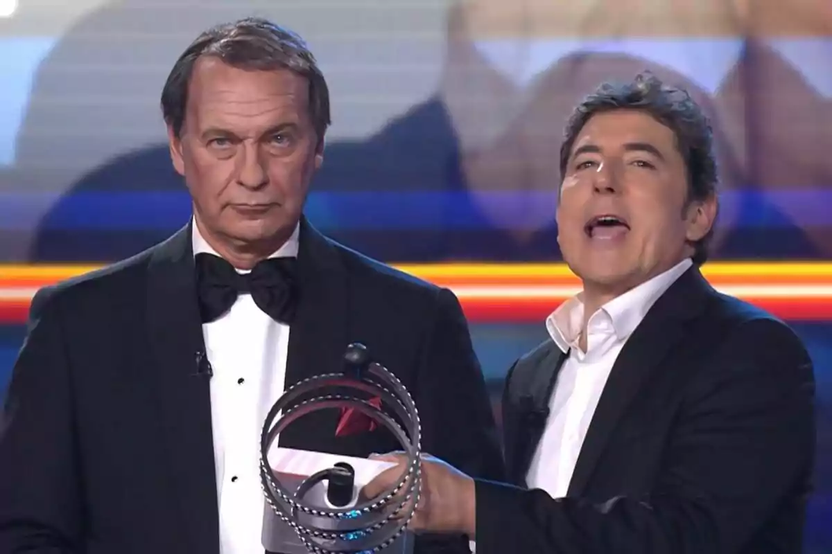 Dos homes en un escenari, Manel Fuentes sosté un objecte circular mentre Bertín Osborne porta un esmòquing i corbata de llaç a Tu Cara Me Suena.