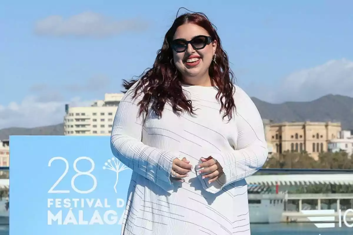 Blanca Martínez amb ulleres de sol i cabell llarg somriu davant d'un cartell del Festival de Màlaga amb un edifici i muntanyes al fons en la presentació de Mariliendre.