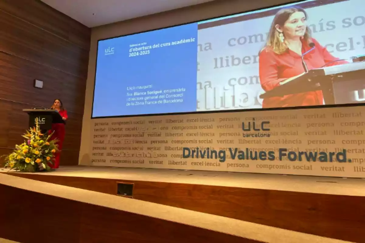 Blanca Sorigué en un podi fent un discurs en un esdeveniment acadèmic en un auditori amb una pantalla gran que mostra la seva imatge i text relacionat amb la inauguració del curs acadèmic 2024-2025 a la ULC.