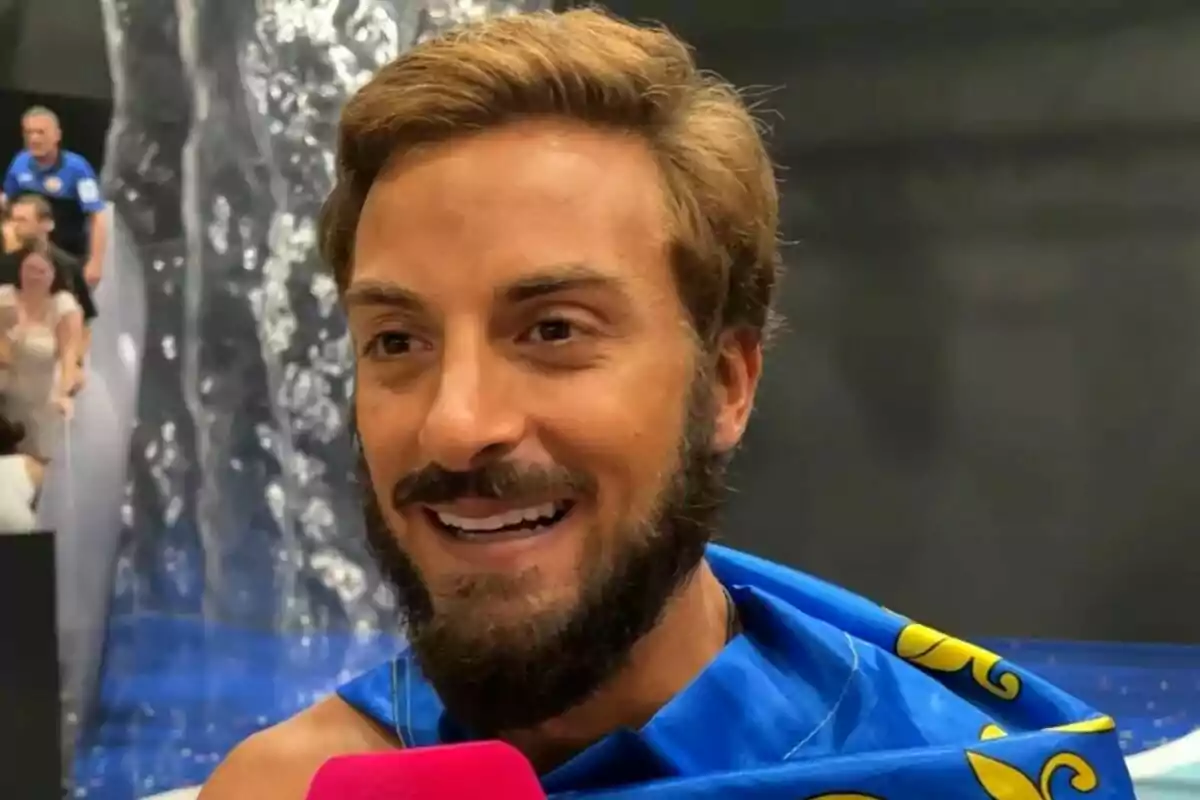 Borja González somrient amb barba i cabell clar embolicat en una bandera de la Comunitat Valenciana blava amb detalls grocs mentre sosté un micròfon rosa al plató de Supervivientes