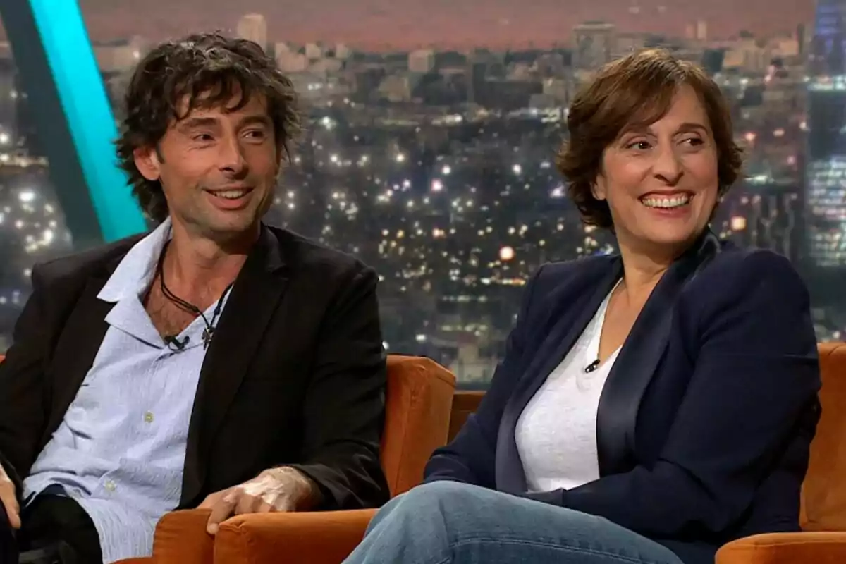 Bruno Oro i Clara Segura assegudes en un sofà durant una entrevista en un programa de televisió, Col·lapse, amb una ciutat il·luminada de fons. Bruno Oro i Clara Segura assegudes en un sofà durant una entrevista en un programa de televisió, Col·lapse, amb una ciutat il·luminada de fons.