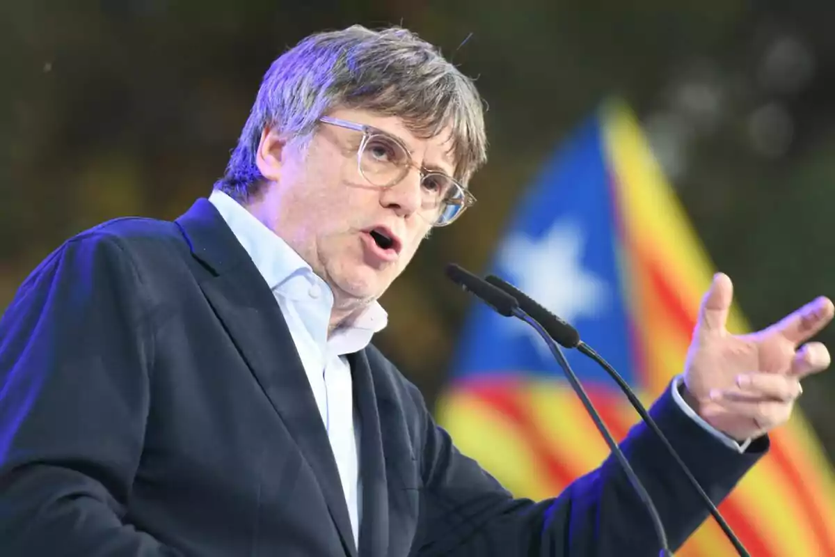 Carles Puigdemont parlant a un micròfon amb una bandera catalana de fons.