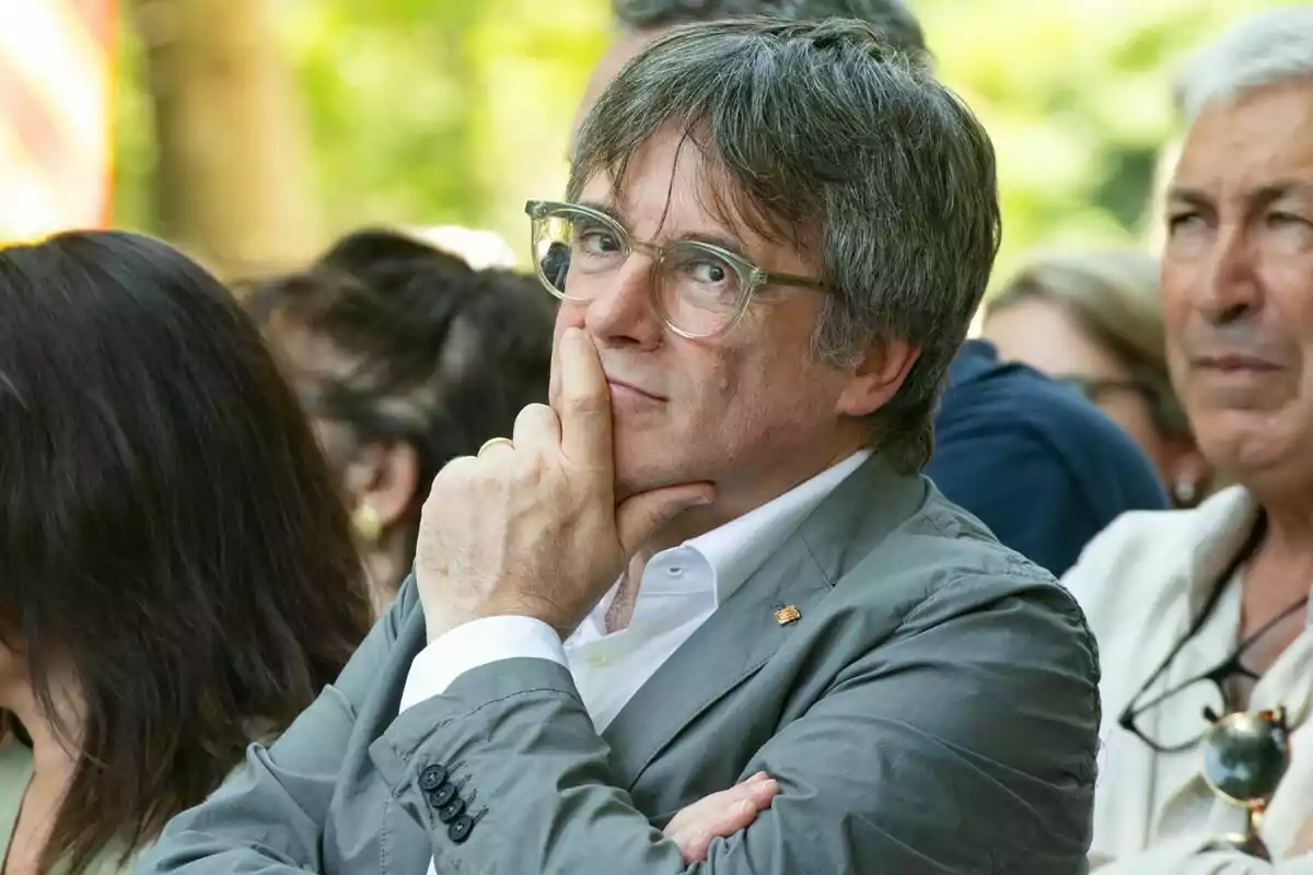 Carles Puigdemont amb jaqueta grisa, amb la mà a la barbeta, sembla pensatiu en un esdeveniment a l'aire lliure.