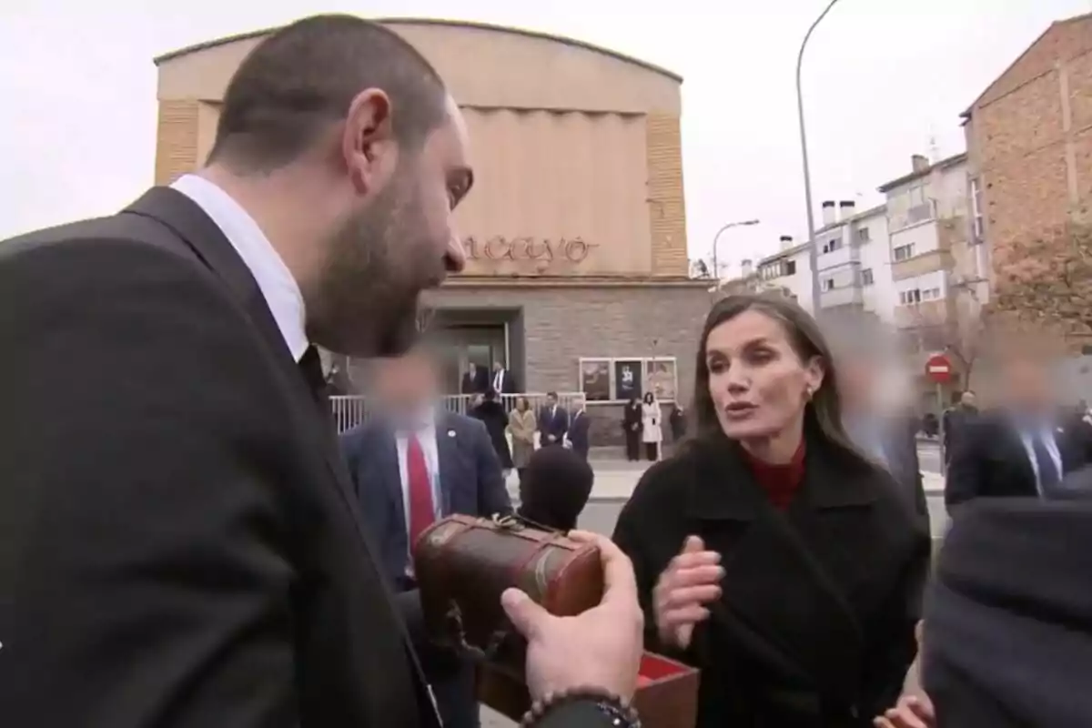 La reina Letizia interactúa con Daniel Fez que sostiene una caja decorativa en un entorno urbano, con un edificio y varias personas de fondo.