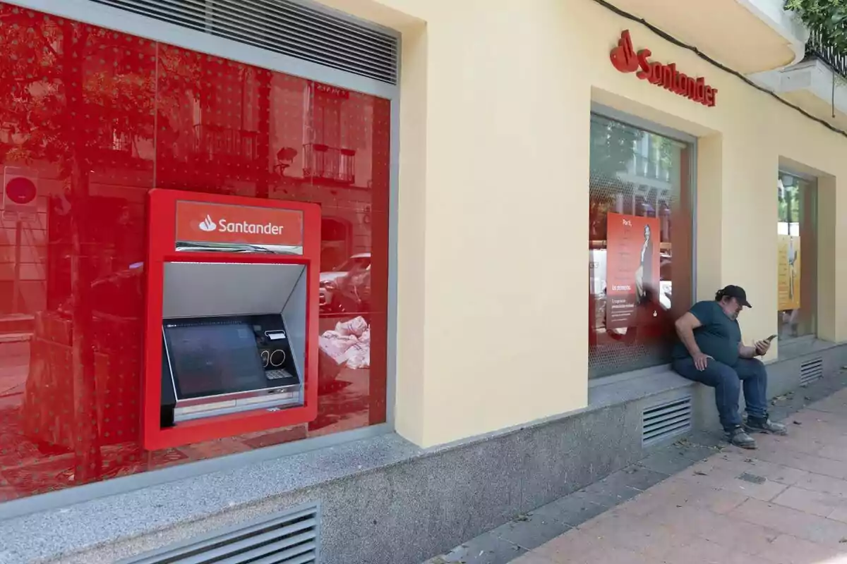 Exterior d'un caixer de Banco Santander amb un home assegut amb gorra i el mòbil a la mà Exterior d'un caixer de Banco Santander amb un home assegut amb gorra i el mòbil a la mà