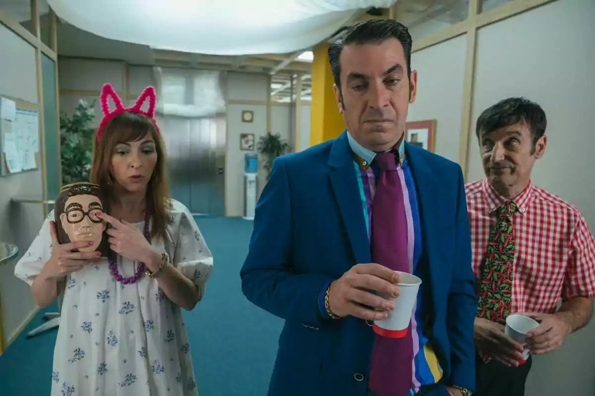 Tres persones en una oficina de la pel·lícula de Camera Café, una dona amb orelles roses sosté una màscara, Arturo Valls amb vestit blau i corbata morada sosté un got i Carlos Chamarro amb camisa de quadres i corbata acolorida els observa.