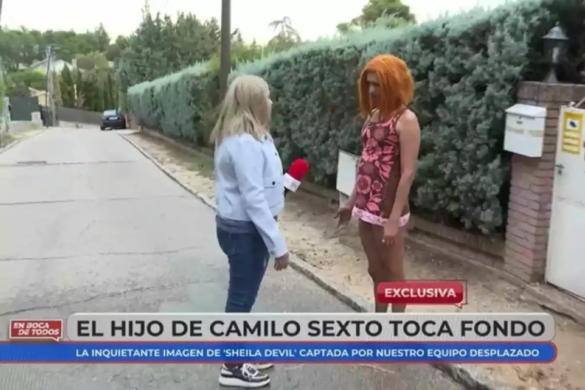 Una reportera d'En Boca de Tots entrevista Camilo Blanes amb perruca taronja i vestit estampat en un carrer residencial, amb una tanca i bústies al fons.