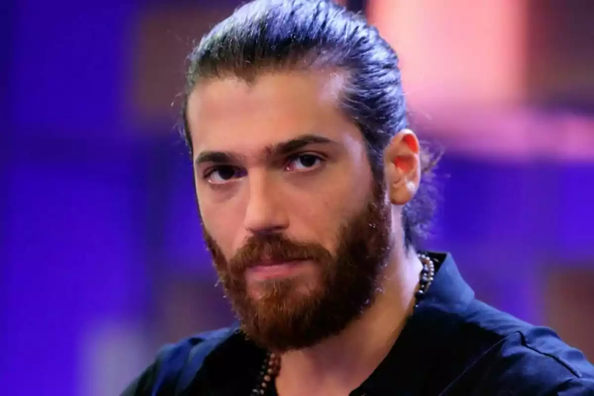 Can Yaman con barba y cabello recogido mira hacia la cámara con un fondo desenfocado en tonos morados y naranjas.