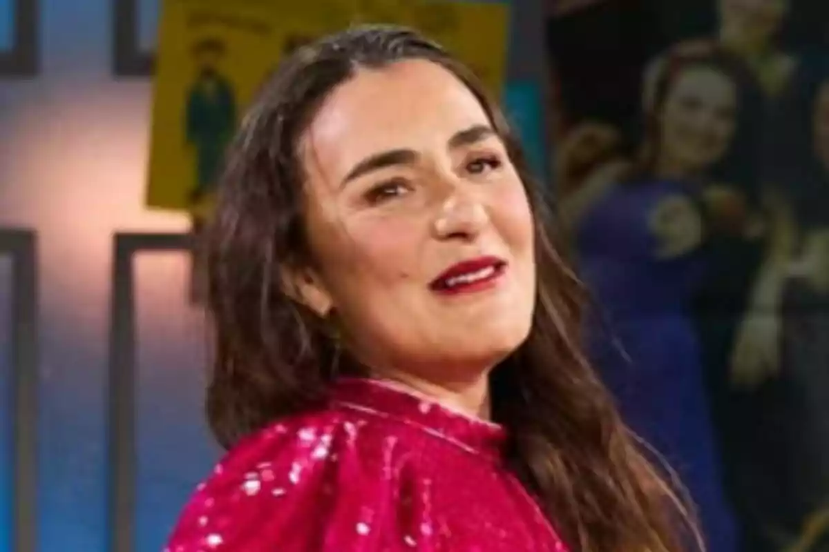 Candela Peña amb cabell llarg i fosc porta un vestit vermell brillant i somriu lleugerament mentre mira cap a un costat en un entorn interior amb llums i cartells de fons a 'La Revuelta'.