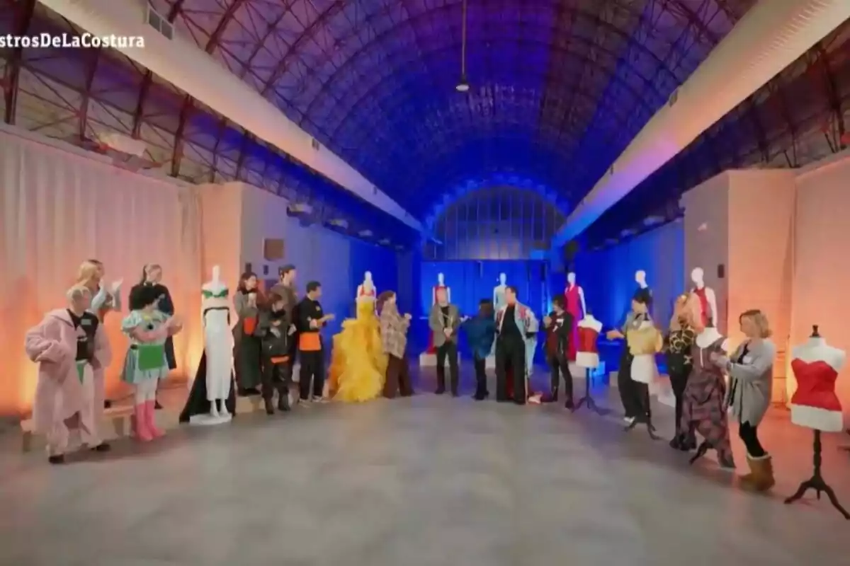 Un grup de persones en un espai ampli amb sostre voltat, envoltats de maniquins amb vestits acolorits i llums de colors al fons a 'Maestros de la Costura Celebrity'. Un grup de persones en un espai ampli amb sostre voltat, envoltats de maniquins amb vestits acolorits i llums de colors al fons a 'Maestros de la Costura Celebrity'.